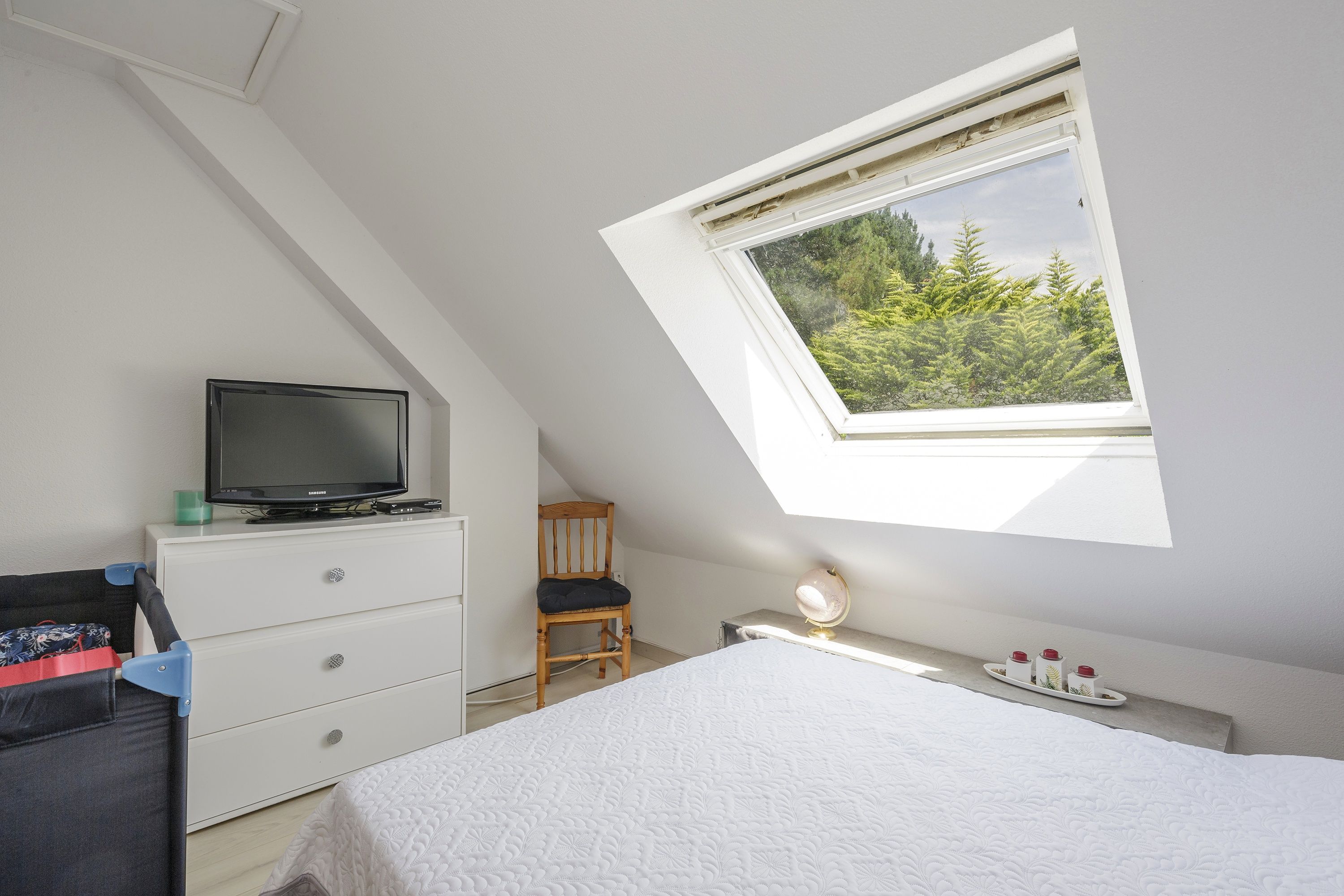 Une chambre avec un plafond en pente, un téléviseur posé sur une commode, un lit recouvert d’une couette blanche, un bureau avec une lampe et une fenêtre donnant sur des arbres.