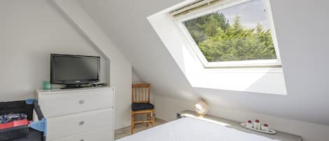 Une chambre avec un plafond en pente, un téléviseur posé sur une commode, un lit recouvert d’une couette blanche, un bureau avec une lampe et une fenêtre donnant sur des arbres.
