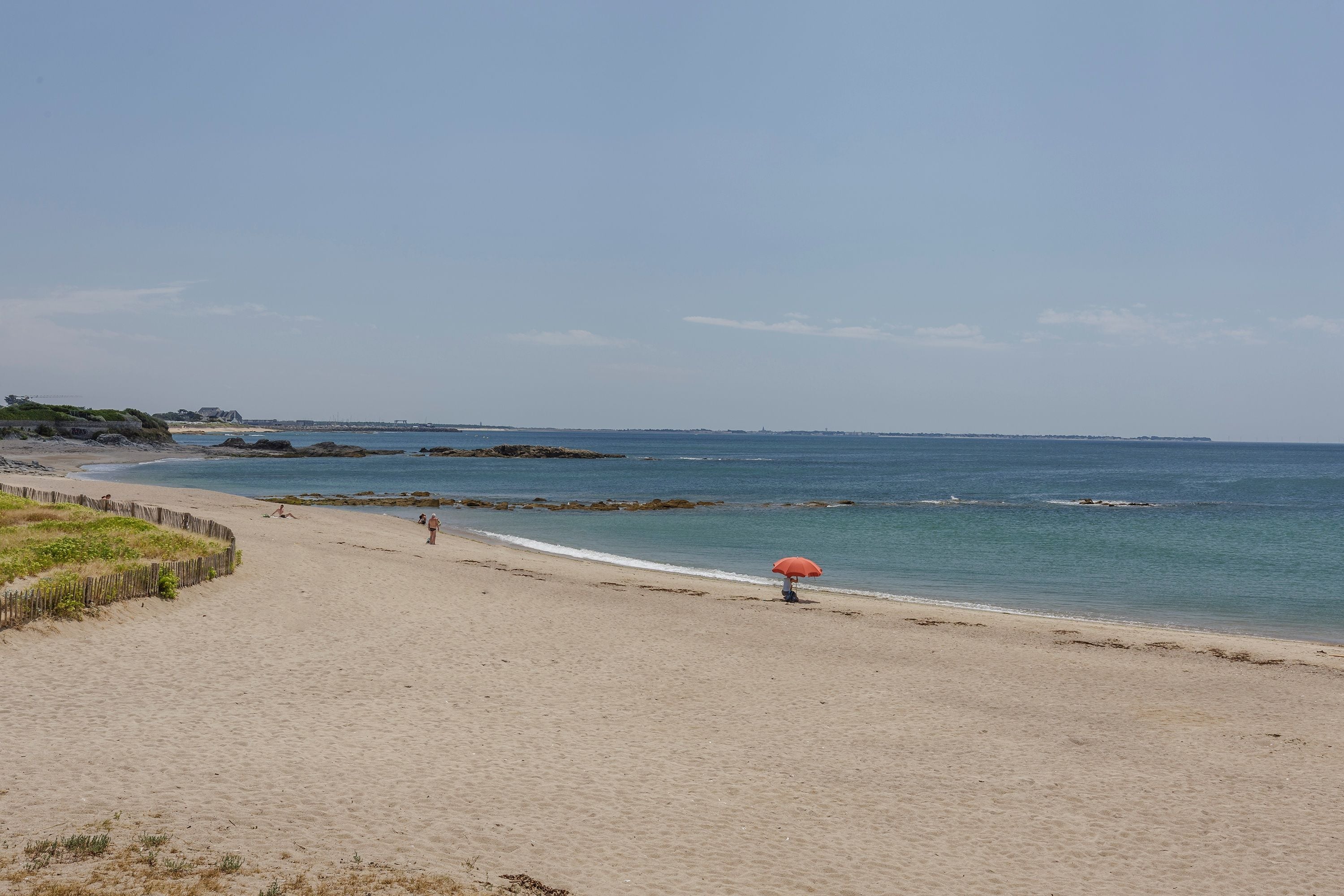 A beach with a wooden fence, calm water, and a person with an umbrella.