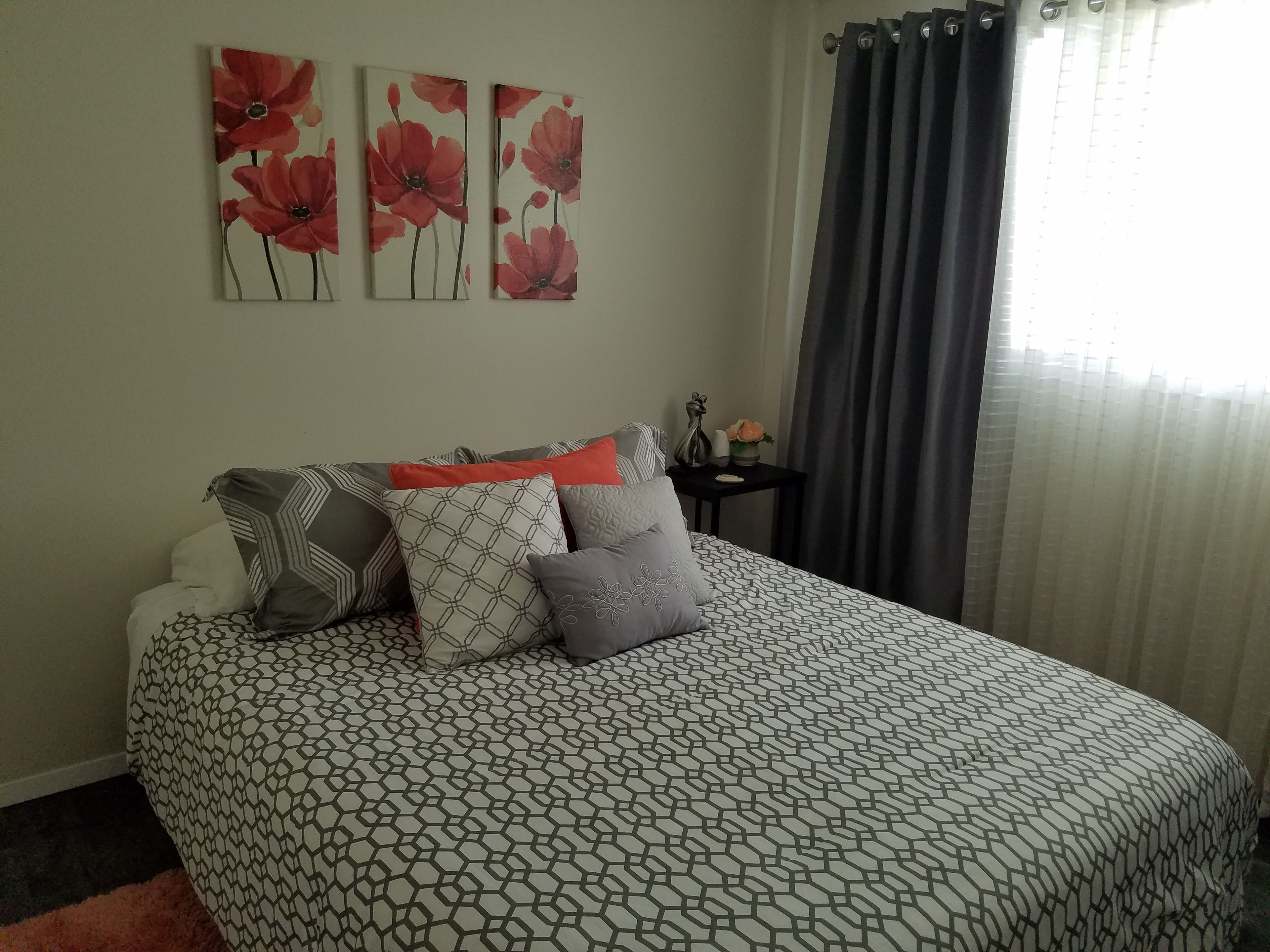 A bedroom with a patterned bedspread, multiple pillows, a nightstand with a vase, and three wall-mounted paintings.