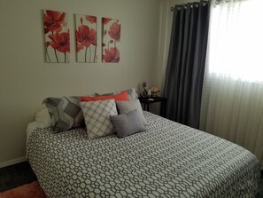Een slaapkamer met een bed met een patroon, meerdere kussens, een nachtkastje met een vaas en drie aan de muur hangende schilderijen.