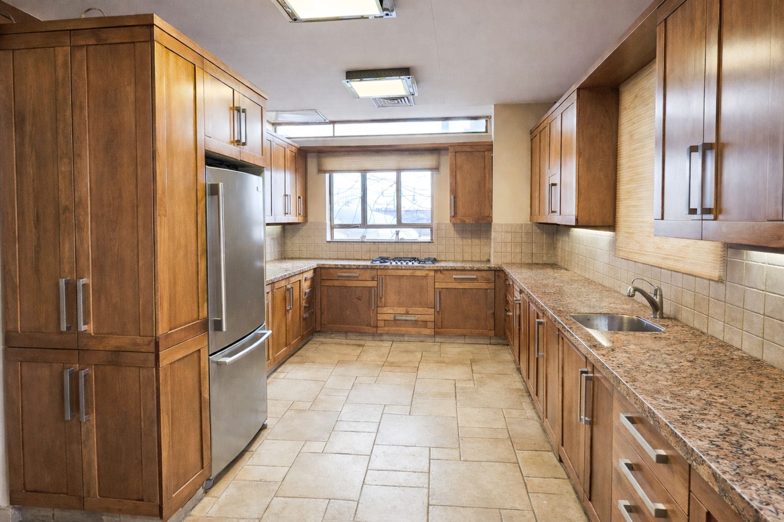A kitchen with wooden cabinets, a stainless steel refrigerator, granite countertops, and a window with blinds.