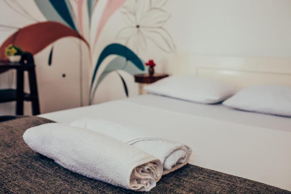 A neatly made bed with a folded white towel on a textured surface, a chair, a small table with a red flower, and a wall with abstract floral patterns.