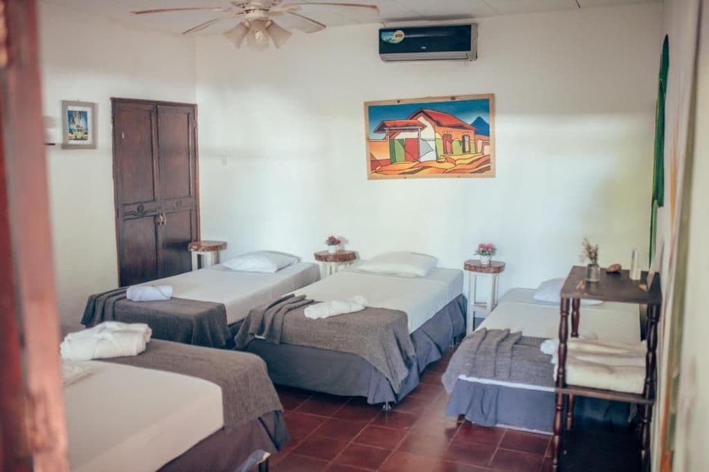 A room with three beds, a ceiling fan, an air conditioning unit, a painting on the wall, and a wooden shelf with towels.