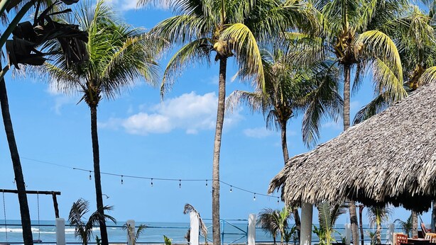 A clear swimming pool with a thatched-roof structure and palm trees in the background. - Casita de Playa Bomalu (Las Peñitas)