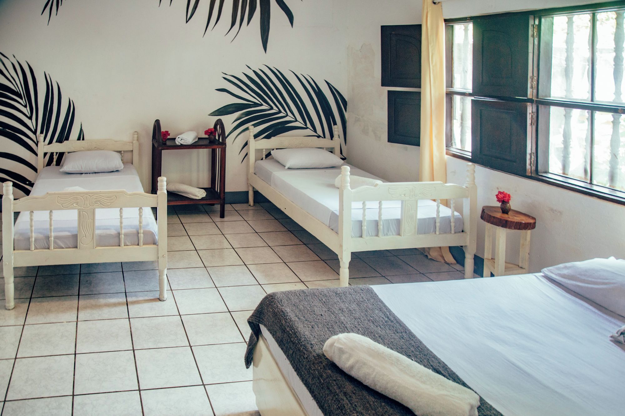 A room with two single beds, a small table, a window with curtains, and a wall with a leaf pattern.
