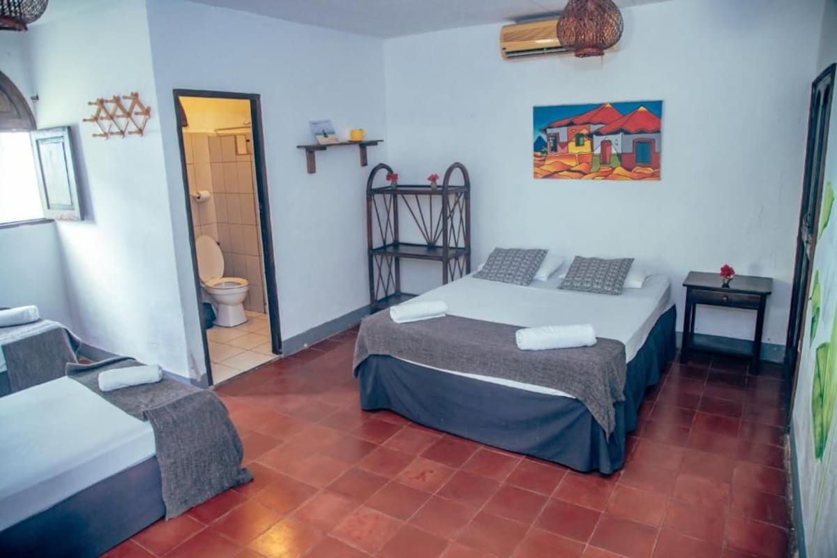 A hotel room with a bed, a small table, a shelf with toiletries, a painting on the wall, and a bathroom with a toilet and shower.