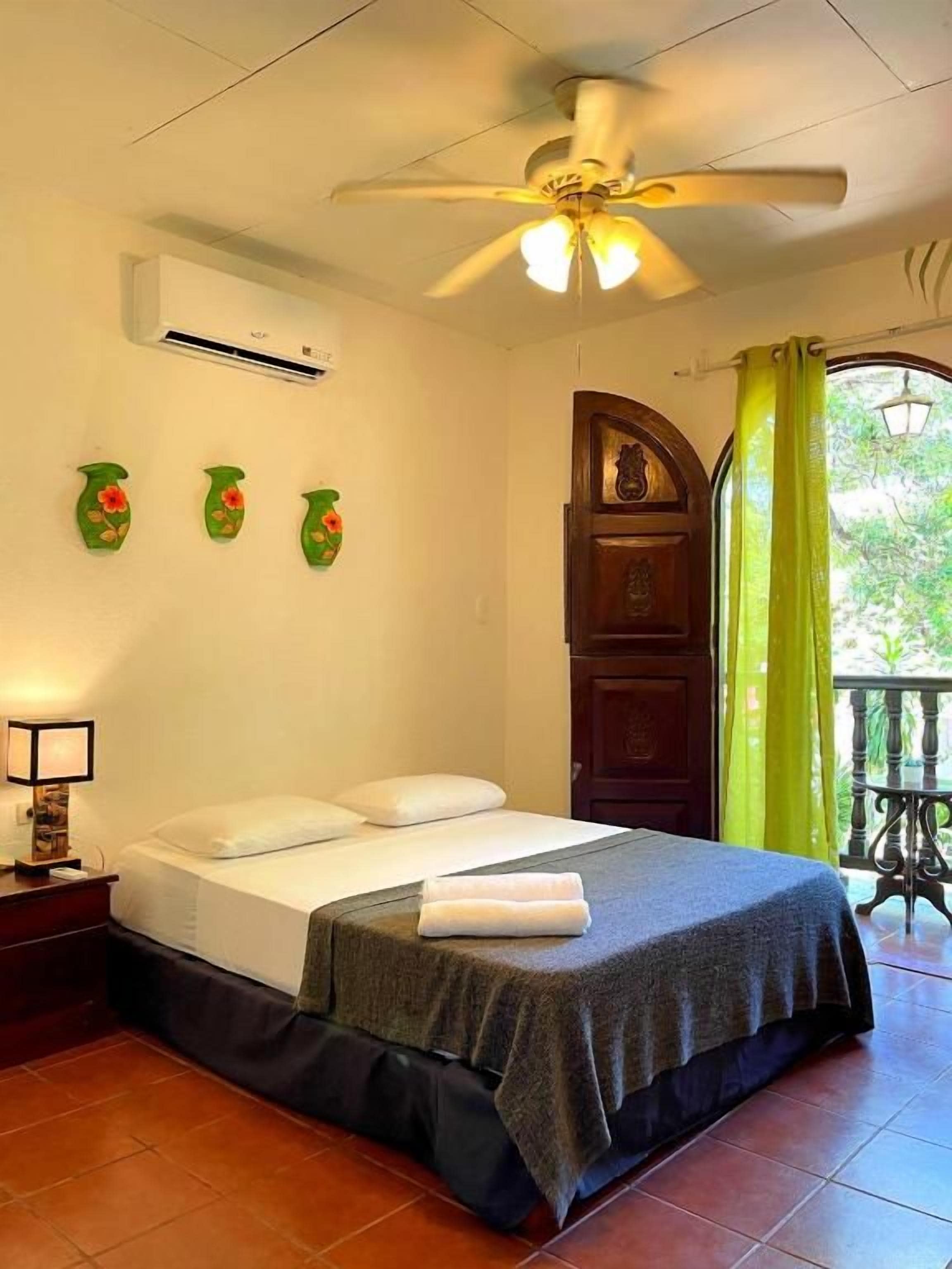 Une chambre à coucher comprenant un lit, un ventilateur de plafond, un appareil de climatisation et une vue sur l’extérieur par une fenêtre munie de rideaux.