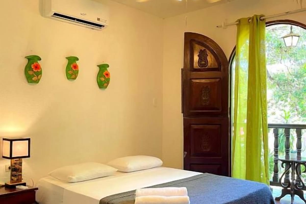 A hotel room with a bed, a ceiling fan, an air conditioning unit, a nightstand with a lamp, and a view of the outdoors through a window with curtains.