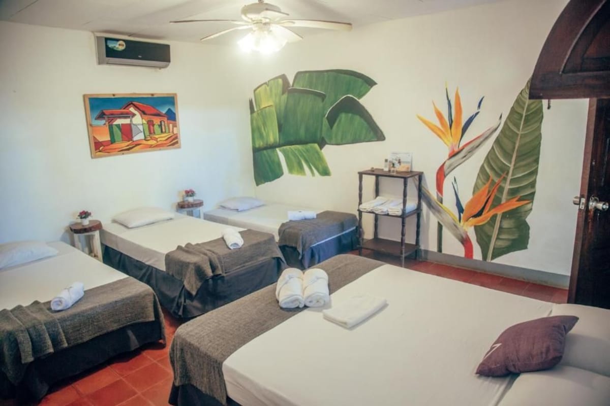 A hotel room with three beds, a ceiling fan, a painting, and a wall mural.