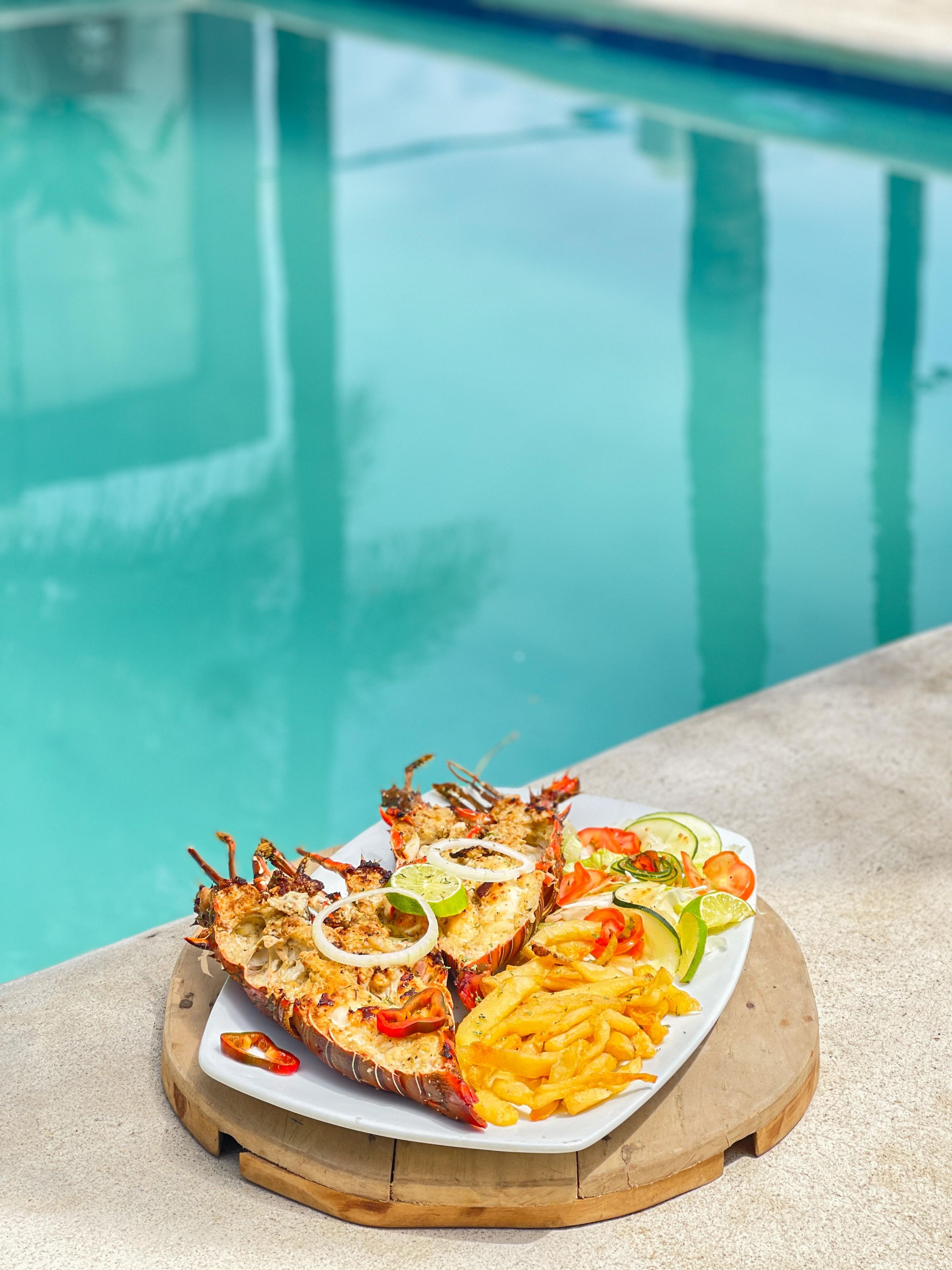 Un plat de fruits de mer servi avec des frites et des légumes sur une planche en bois, près d’une piscine.