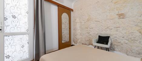 A bedroom with a stone wall, a bed with a beige cover, a chair with a black cushion, and a door with a floral pattern.