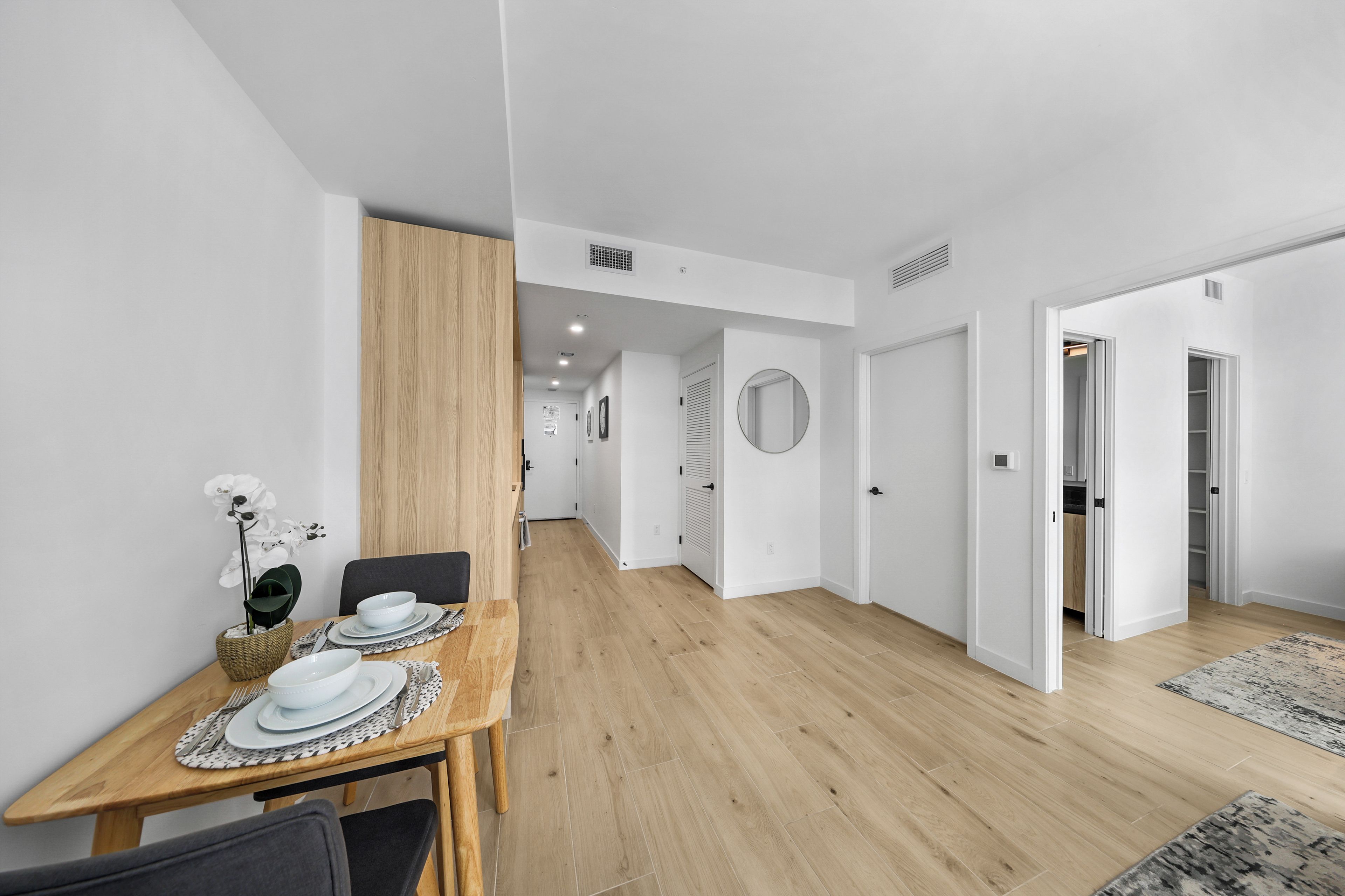 A modern interior with a dining area featuring a wooden table set for four, a hallway leading to a bedroom, and a bathroom with a shower and toilet.