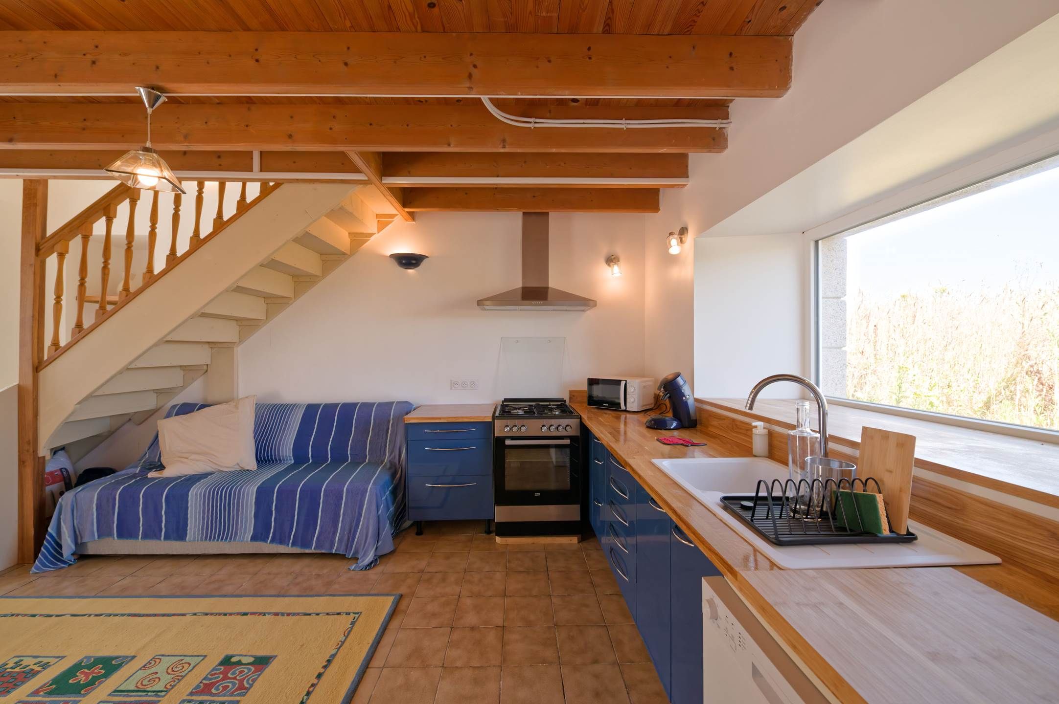 Een compacte keuken met een fornuis, oven, magnetron en spoelbak. Er is een bed met gestreepte beddengoed en een trap die naar een hoger gelegen niveau leidt.