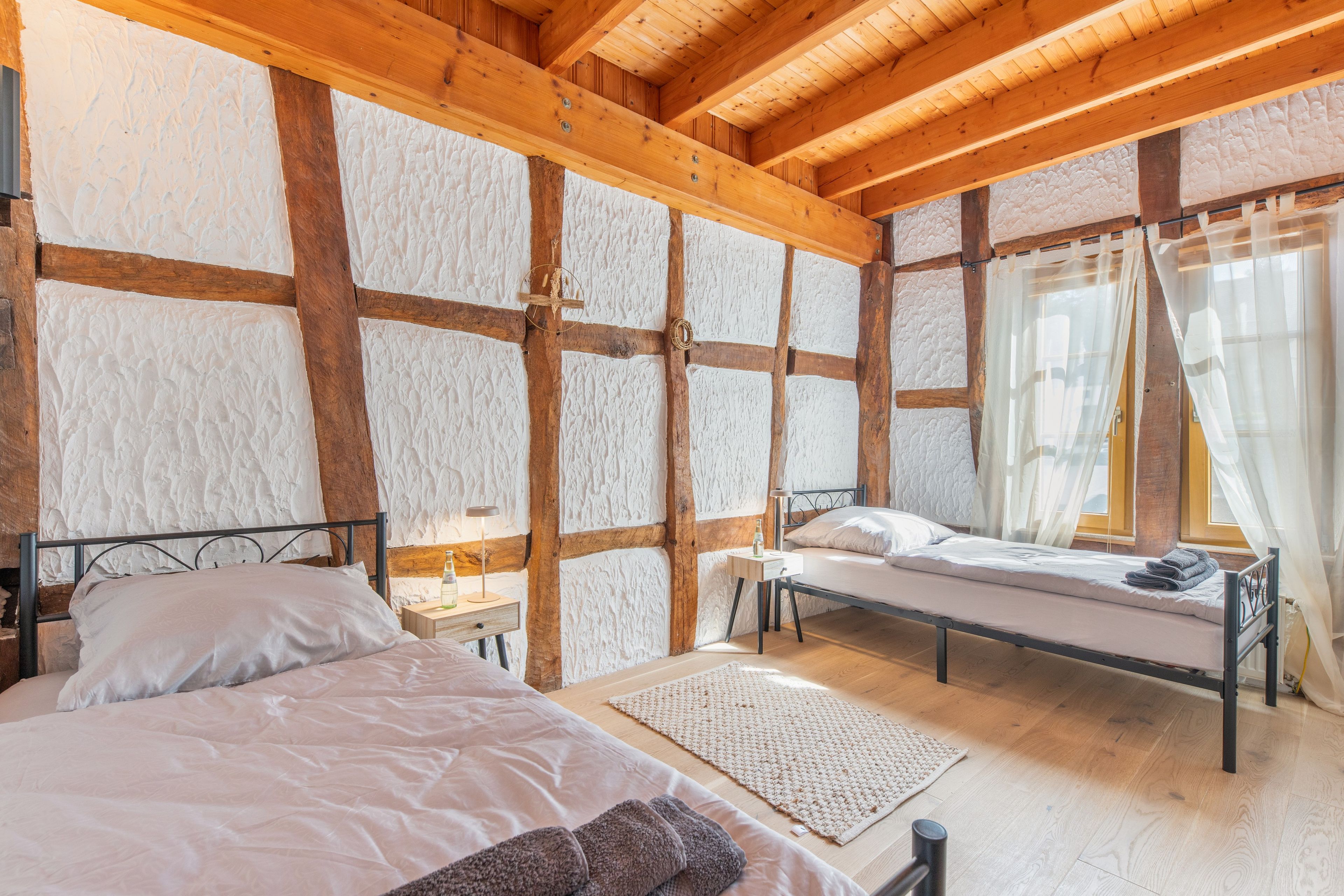 Een slaapkamer met een houten plafond, een bed met witte lakens, een nachtkastje met een lamp en een raam met gordijnen.