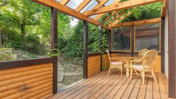 A wooden deck with a glass ceiling, surrounded by greenery, and furnished with wicker chairs and a table.