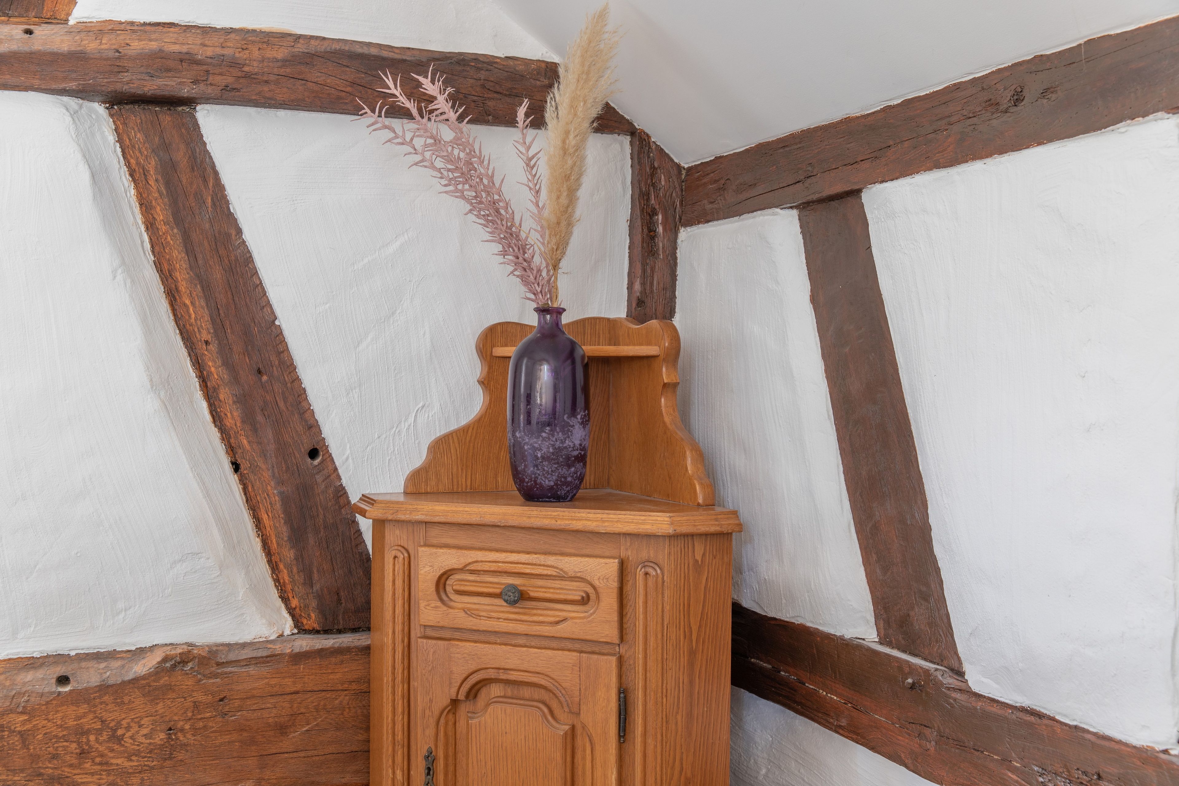 A wooden cabinet with a vase containing dried plants, set against a white wall with exposed wooden beams.