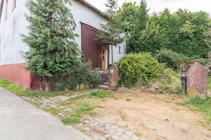 A house with a stone pathway leading to a wooden door, surrounded by greenery and a stone wall.