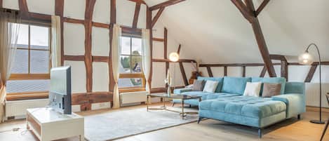 A loft-style room with exposed wooden beams, a large window with curtains, a blue sofa, a standing lamp, and a television on a white stand.