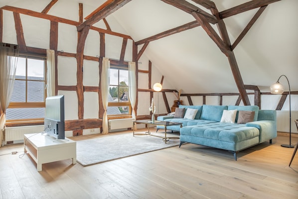 A loft-style room with exposed wooden beams, a large window with curtains, a blue sofa, a standing lamp, and a television on a white stand.