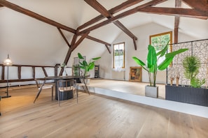 A spacious loft with exposed wooden beams, a dining area with a table and chairs, and large green plants.