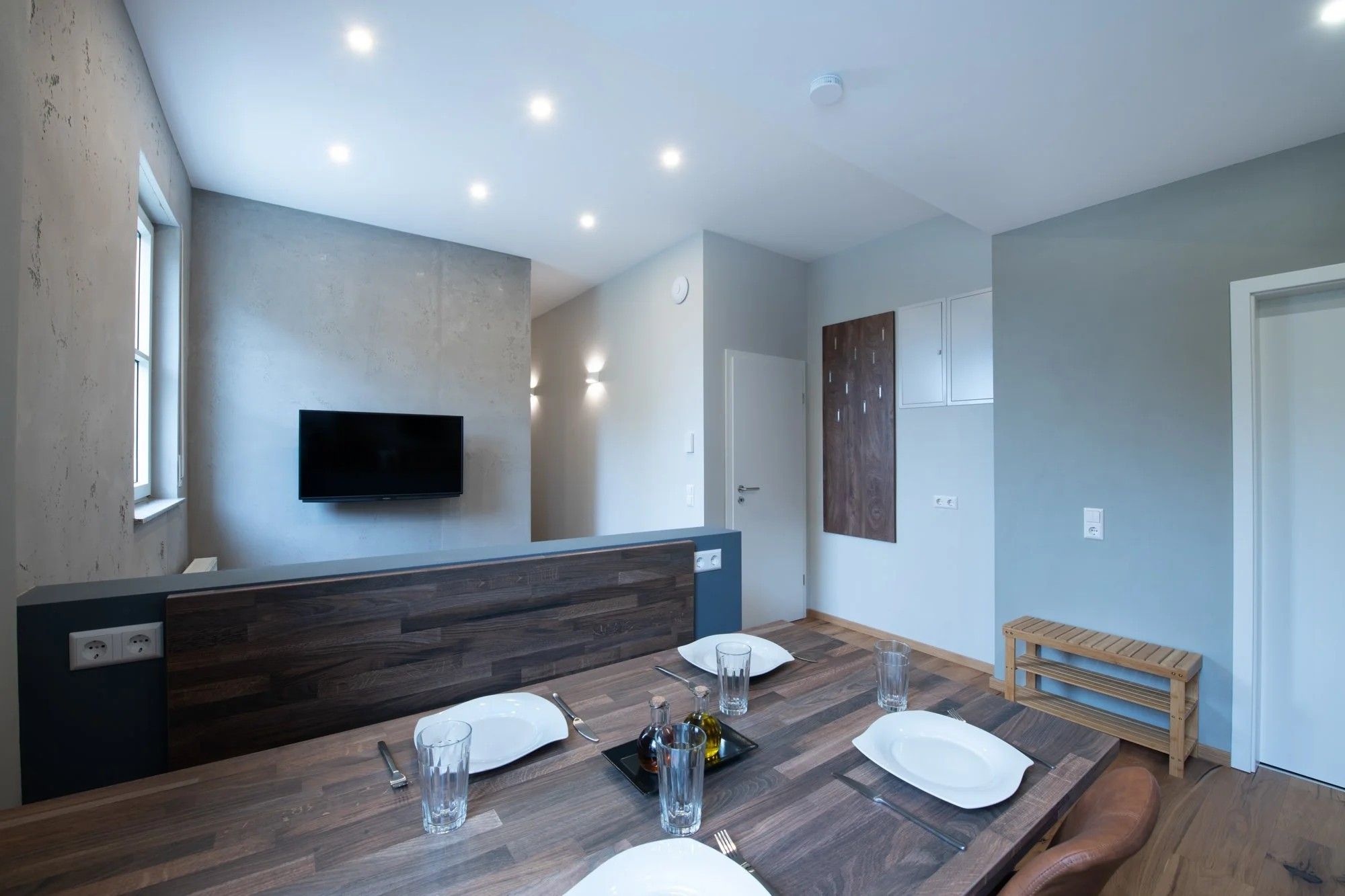 A modern dining area with a wooden table set for two, a flat-screen TV mounted on the wall, and a bench in the corner.