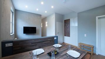 A modern dining area with a wooden table set for two, a flat-screen TV mounted on the wall, and a bench in the corner.