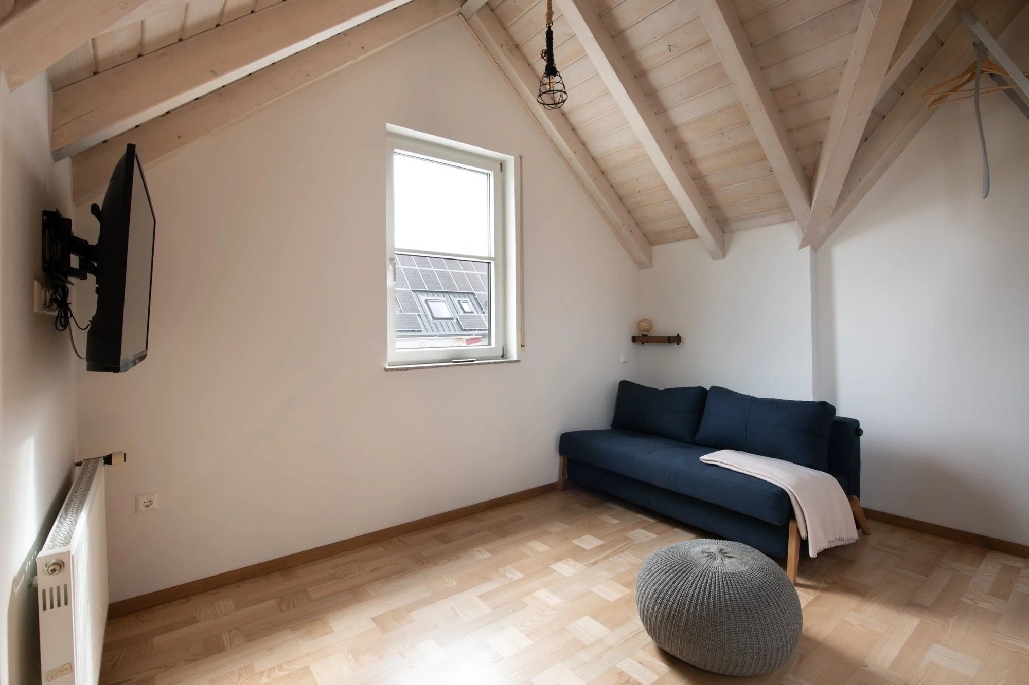 Ein Zimmer mit schrägen Wänden, einem Fenster mit Solarzellen, einem blauen Sofa, einem Heizkörper und einer Pendelleuchte.