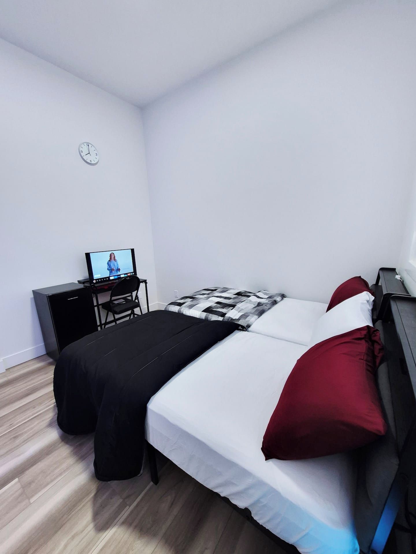 Quarto pequeno e modesto com uma cama, uma escrivaninha e uma cadeira. Há um relógio na parede.