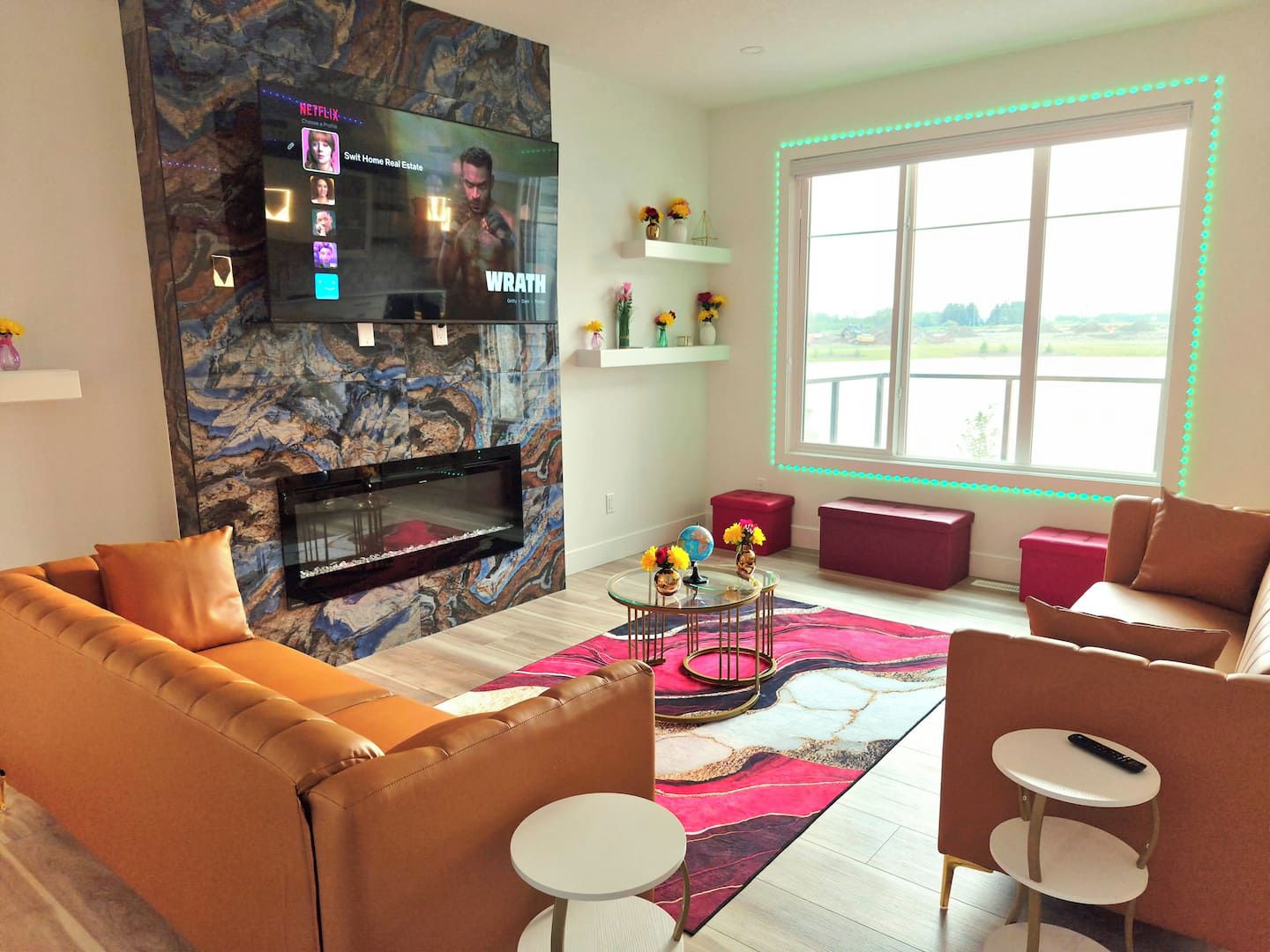 Uma sala de estar moderna com televisão de tela plana fixada em uma parede de mármore, lareira abaixo, janela grande com vista e um espaço de estar aconchegante com um banquinho vermelho e um sofá rosa.