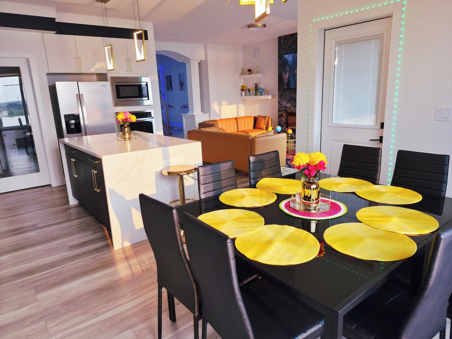 Una cocina moderna con una isla central, un área de comedor con manteles individuales amarillos y un espacio de sala con sofá y mesa de centro.