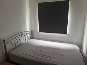 A single bed with a metal frame and white bedding in a room with white walls and a window with a black blind. - Apartment Sleeps 4 With Parking (London)