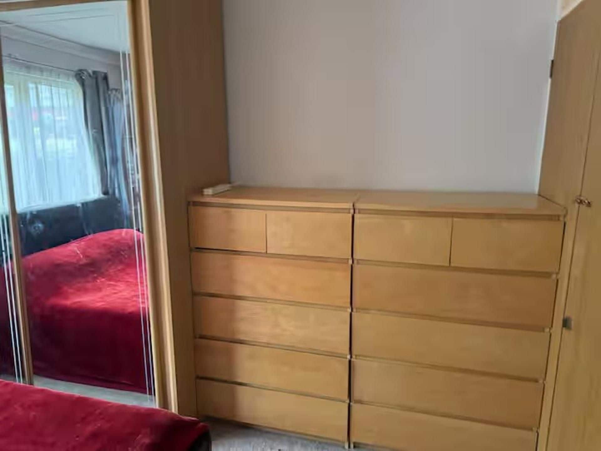 Ein Zimmer mit einem Holfschrank, einer roten Tagesdecke und einem Fenster mit Vorhängen.