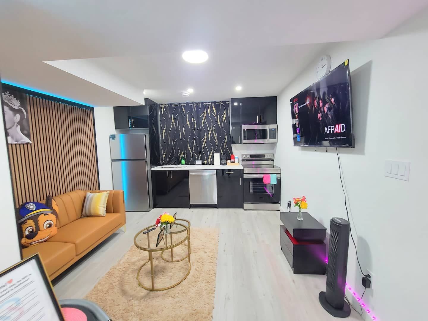 A modern living room with a sofa, a coffee table with flowers, a TV mounted on the wall, and a kitchen area with appliances.