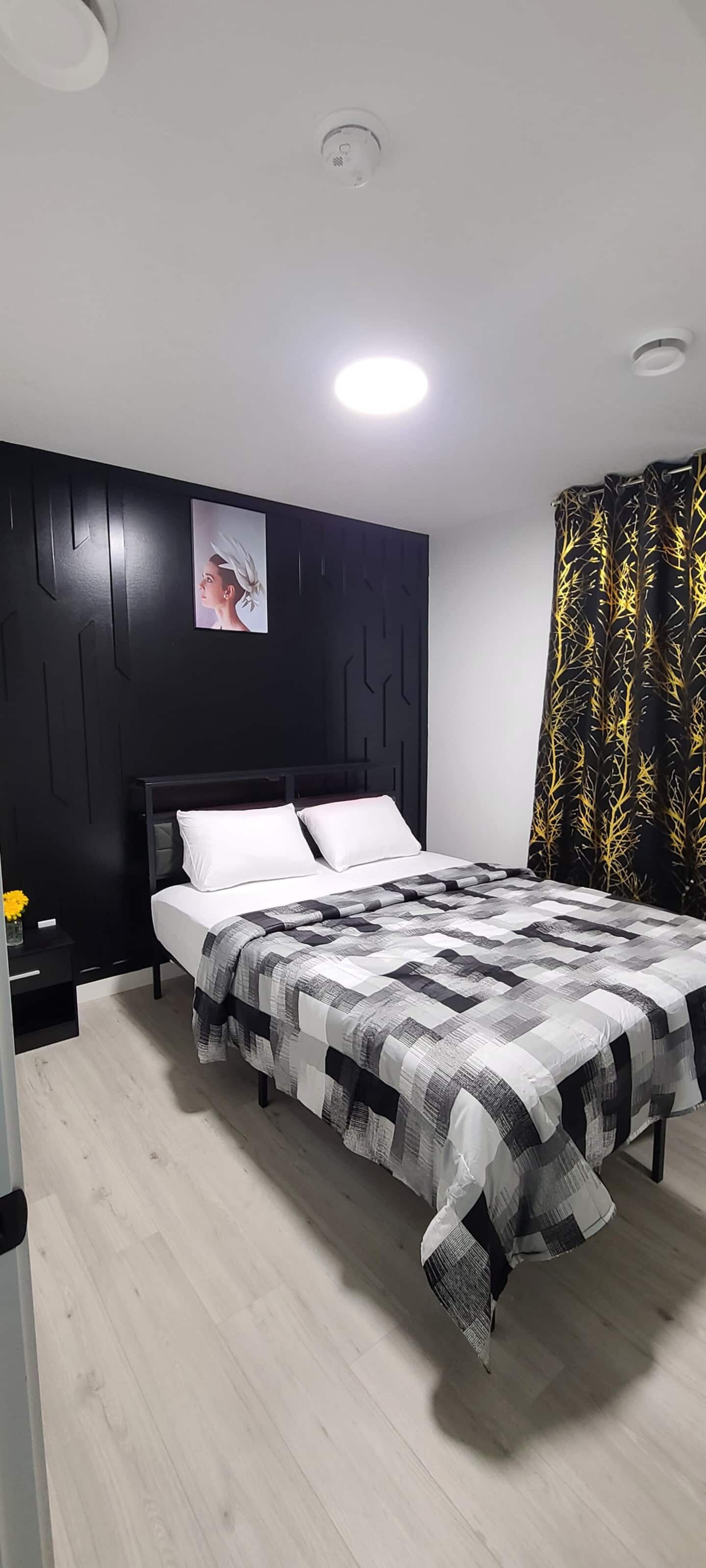 Un dormitorio moderno con cabecera negra, cama con colcha a cuadros, mesita de noche y un cuadro colgado en la pared.