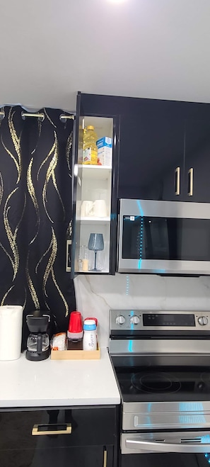 A kitchen with a microwave, oven, and a cabinet with a glass door containing a bottle and a box. - SwitHome Properties Glen Cozy Suite Near Yeg Airport (Edmonton)
