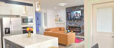 Una cocina moderna con una isla central, un comedor con mesa y sillas, y una sala de estar con sofá y televisor montado en la pared.