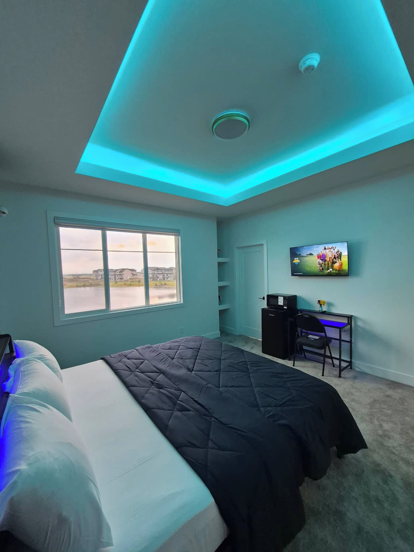 A bedroom with a bed, a desk, a television, and a window with a view of buildings and a body of water.