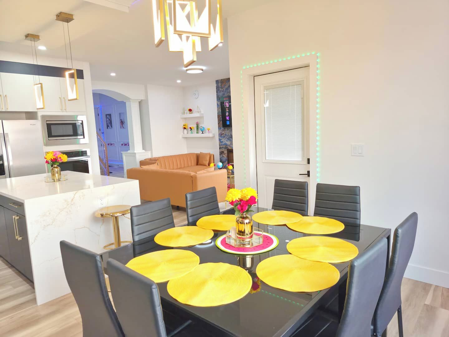 A modern kitchen with a central island, a dining area with a black table and yellow placemats, and a living room with a brown sofa.