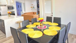 Una cucina moderna con isola centrale, una zona pranzo con tavolo nero e tovagliette gialle, e un soggiorno con divano marrone.