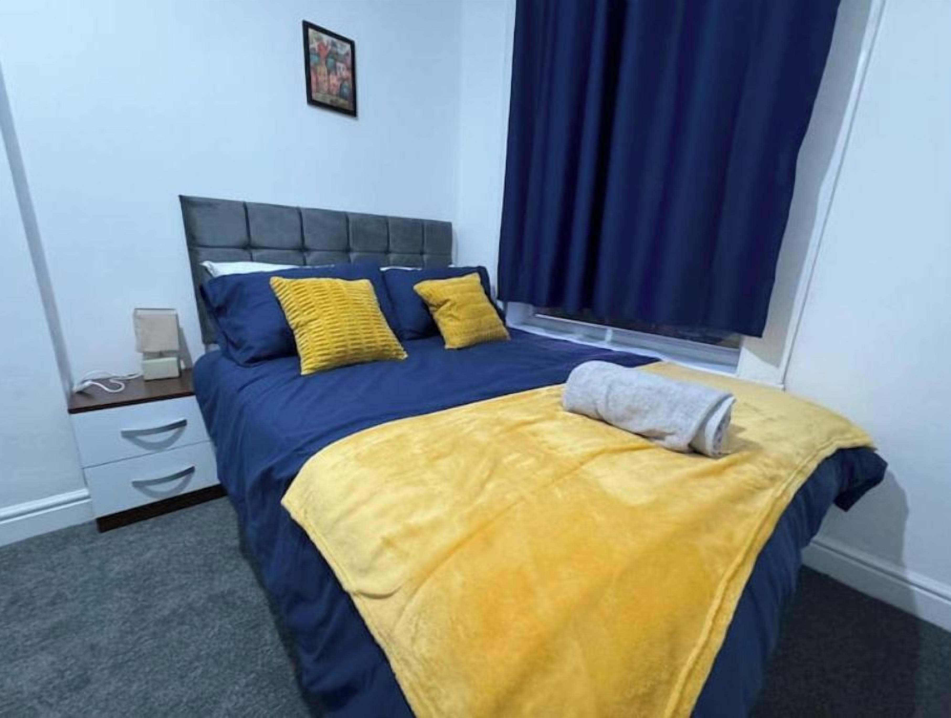 Ein ordentlich bezogenes Bett mit blauer und gelber Bettwäsche, ein Nachttisch mit Lampe und ein Fenster mit Vorhängen.