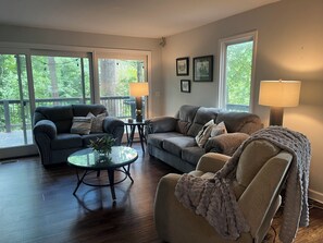 A living room with two sofas, a coffee table, and a lamp. There are large windows with a view of trees. - Clemson Game-Day Retreat - 4BR, Sleeps 8, 5 Min to Death Valley (Clemson)