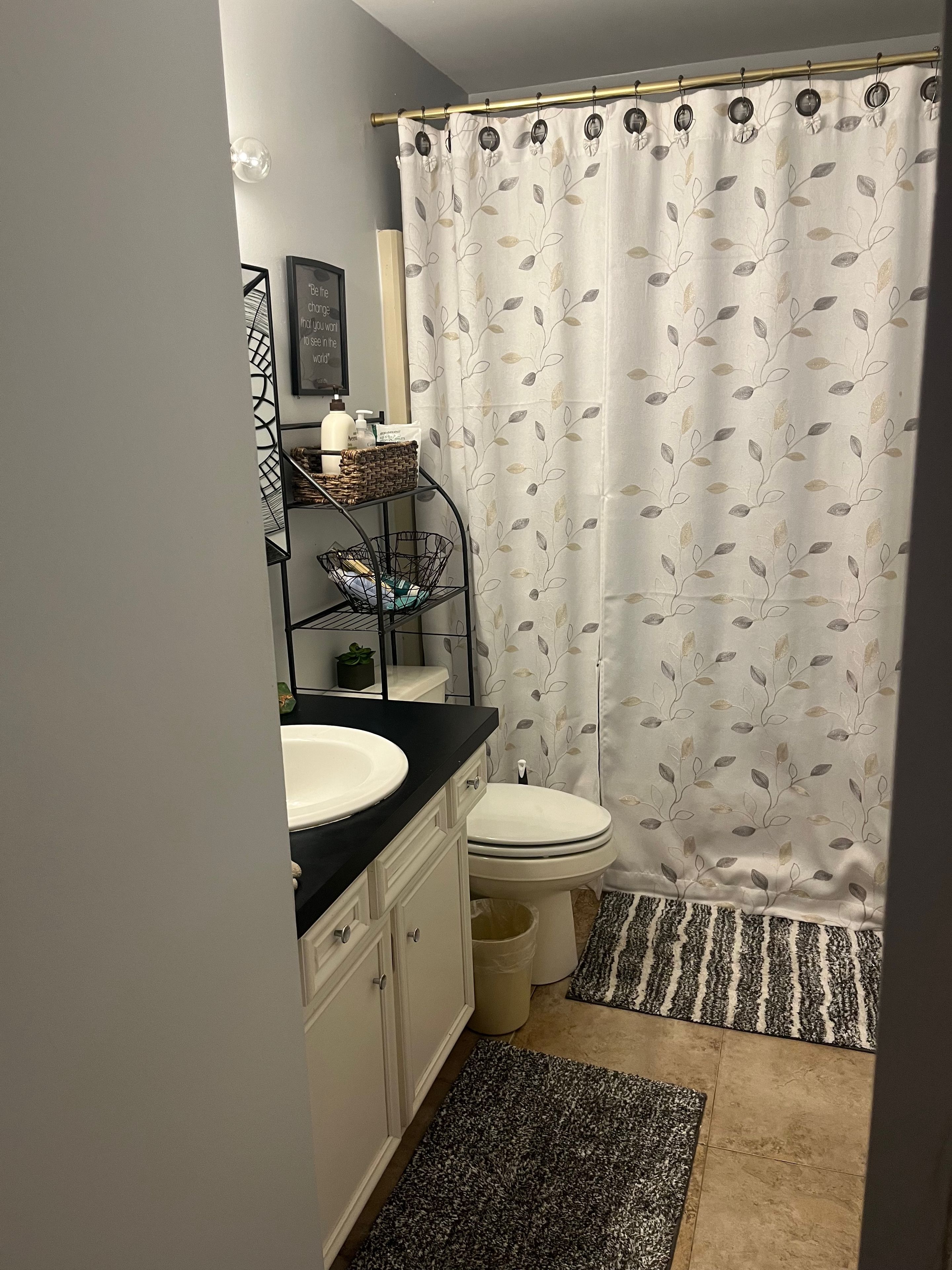 A bathroom with a toilet, a sink, a shower curtain with a leaf pattern, a small shelf with toiletries, and a rug on the floor.