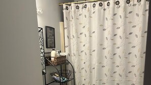 Et badeværelse med toilet, håndvask og bruseforhæng. Der er en hylde med toiletartikler og en lille plante.