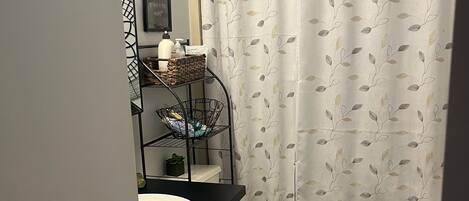 A bathroom with a toilet, a sink, a shower curtain with a leaf pattern, a small shelf with toiletries, and a rug on the floor.