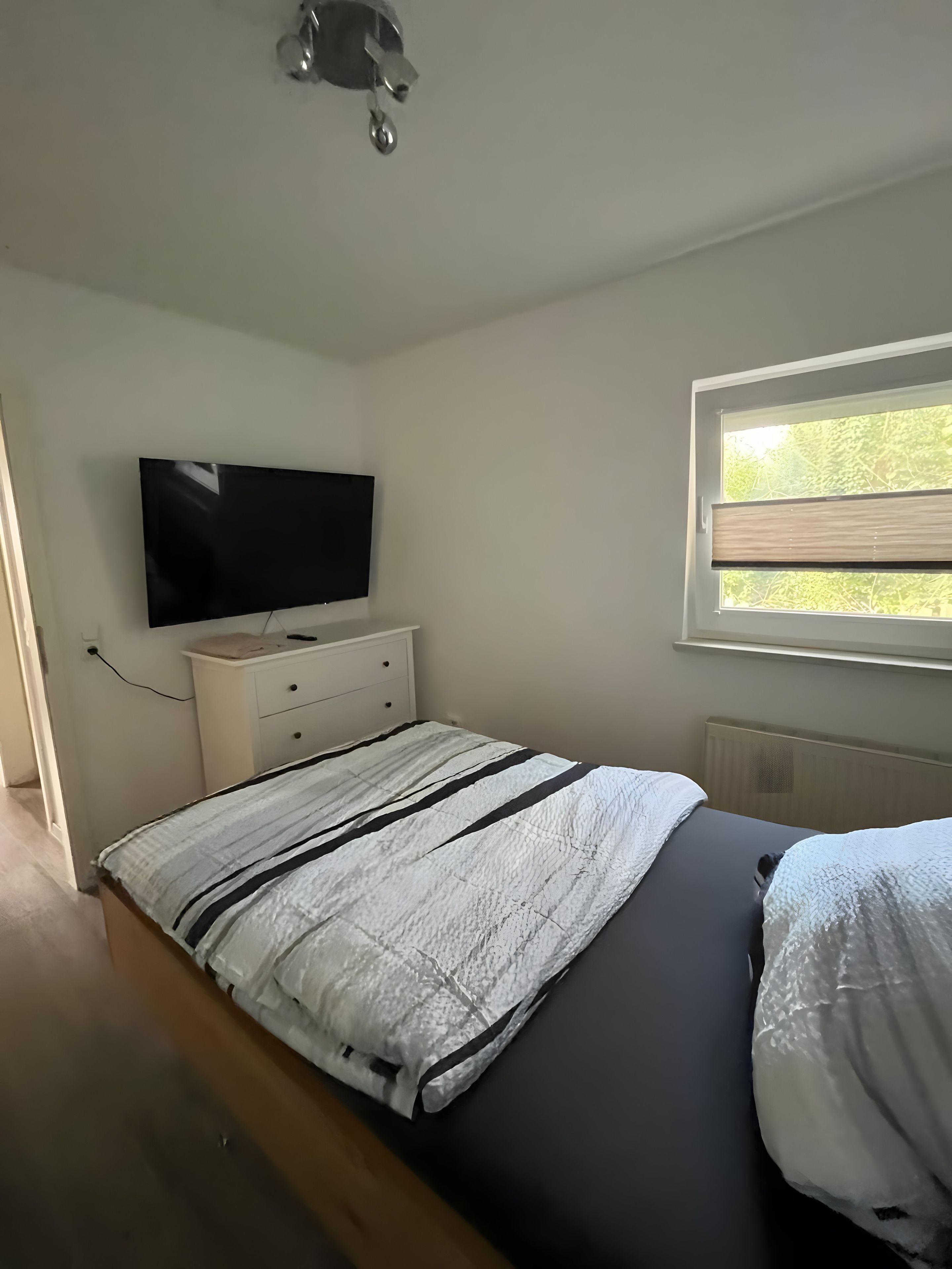 A bedroom with a bed, a television on a dresser, a window with blinds, and a radiator.