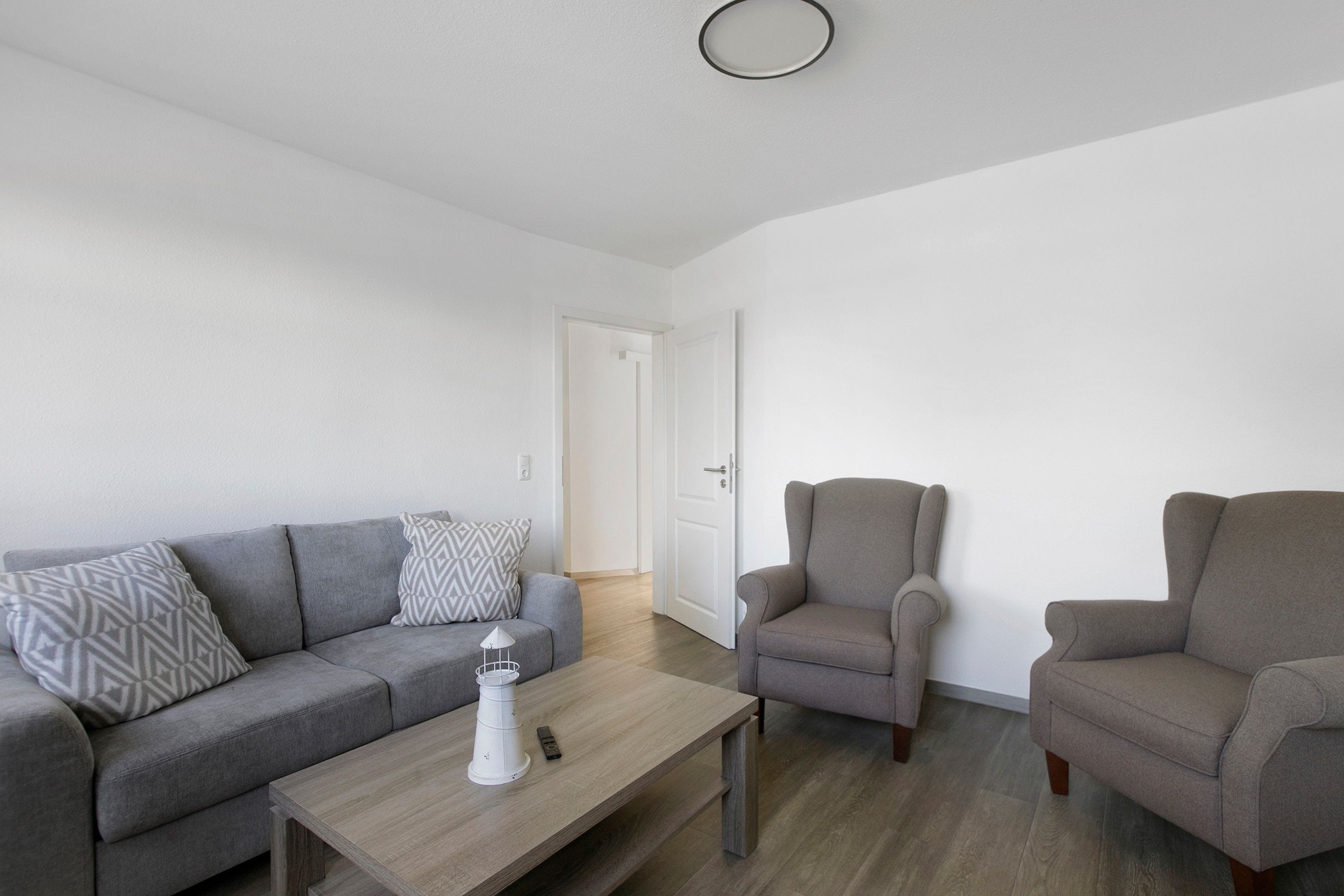 Un salon moderne avec un canapé gris, deux fauteuils, une table basse et une porte blanche en arrière-plan.