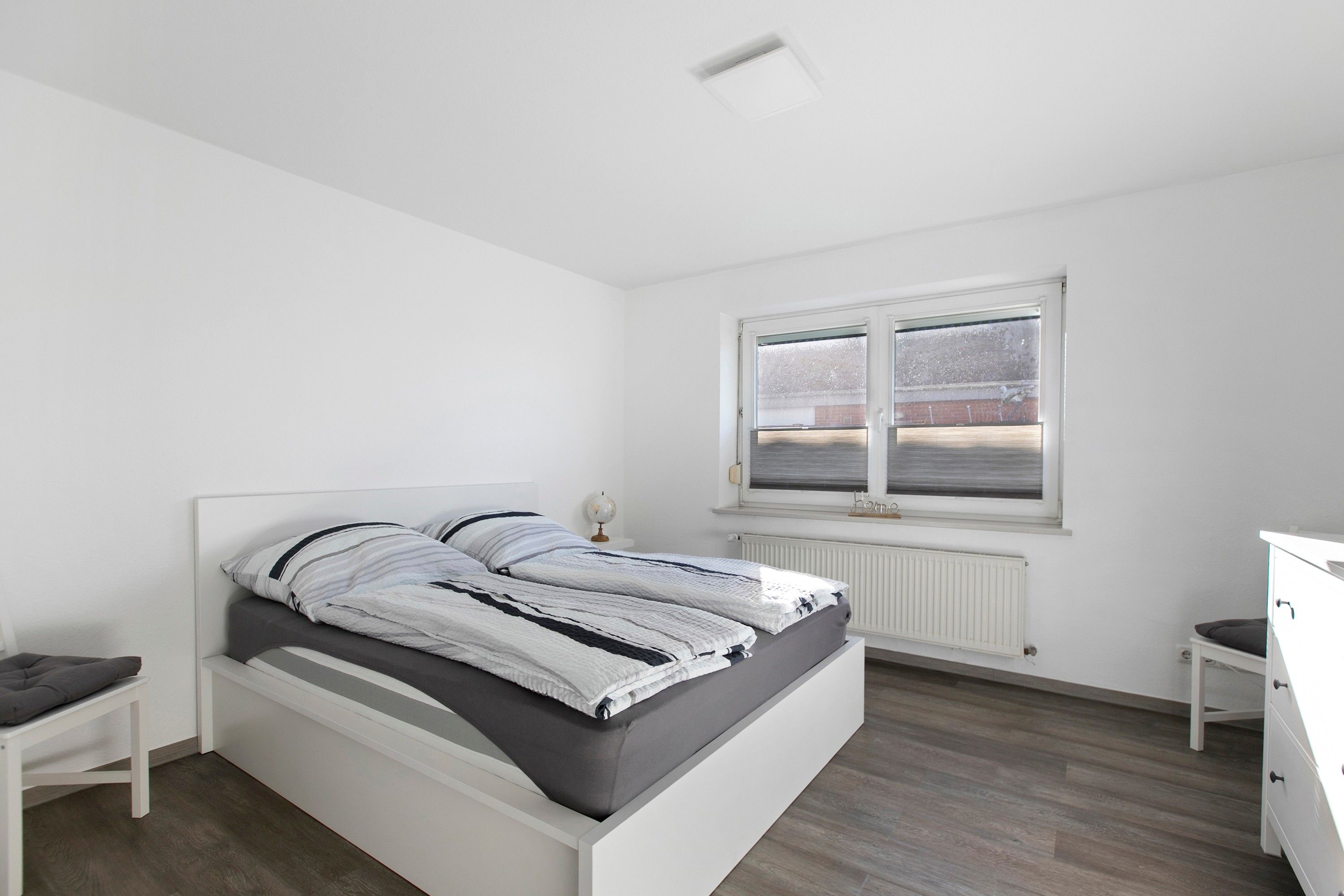 Une chambre moderne avec un lit simple, une table de chevet, une commode et une fenêtre donnant sur un immeuble en briques.