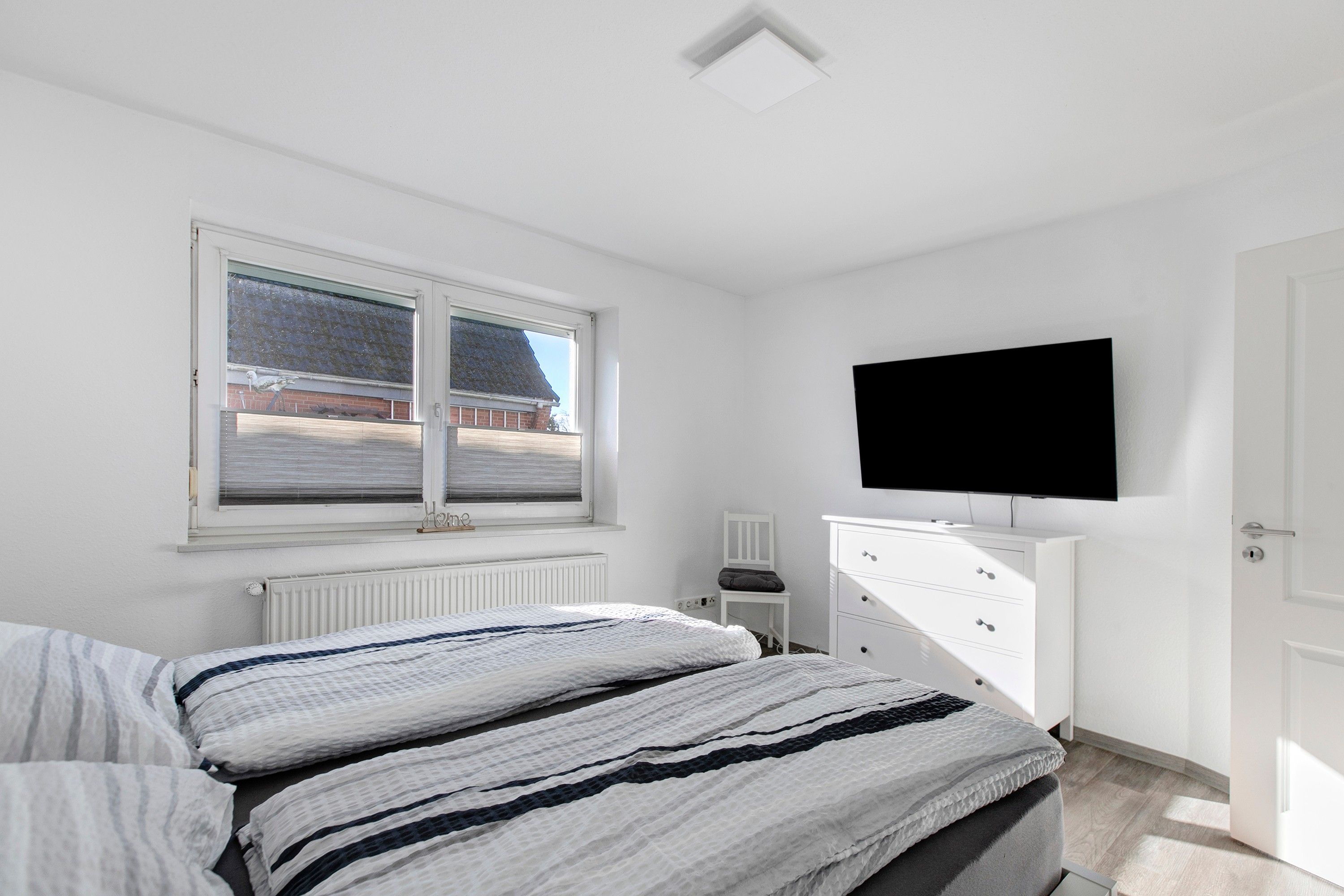 Een slaapkamer met een bed, een aan de muur bevestigde televisie, een raam met zonwering en een radiator.