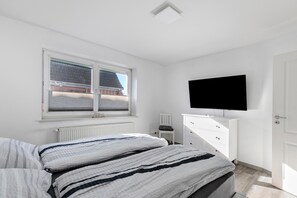 Un dormitorio con cama, televisor montado en la pared, ventana con persianas y radiador.